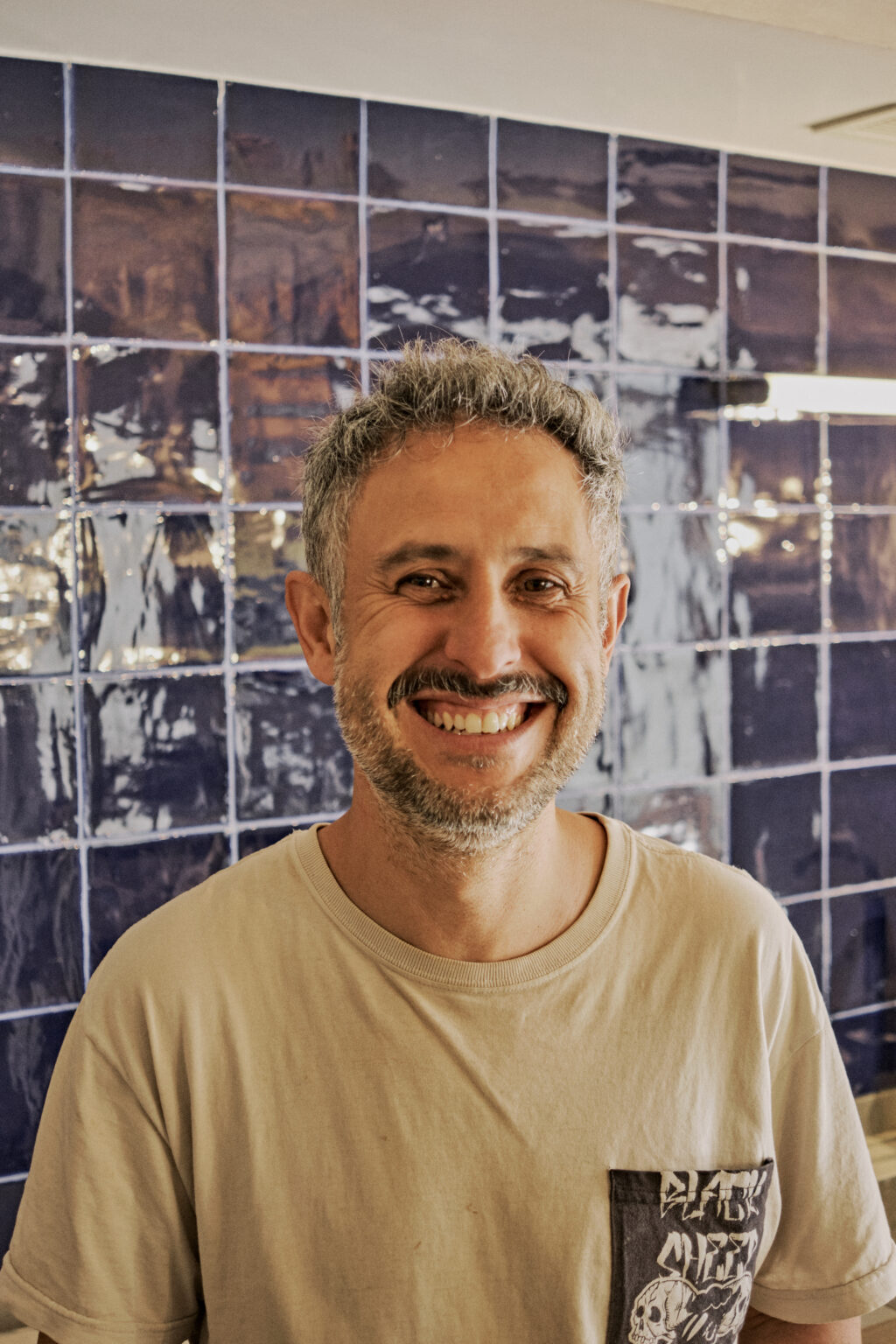 Smiling man with gray hair and a beard, wearing a light shirt, against a blue tiled background.