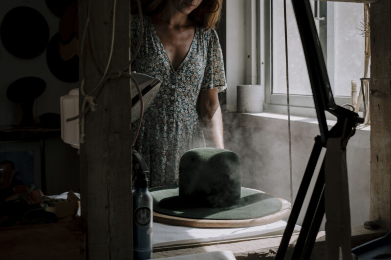 A woman in a floral dress steams a green hat in a workshop, with sunlight streaming through a window.