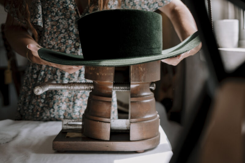 A person adjusts a green felt hat on a wooden hat block, surrounded by soft natural light.