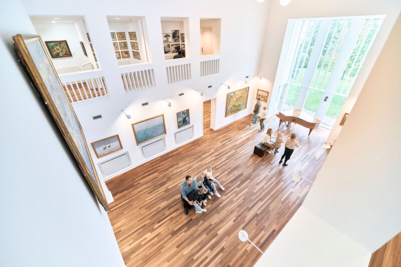 Bright interior of Johannes Larsen Museet, featuring art on walls and visitors seated on benches. Large windows allow natu...