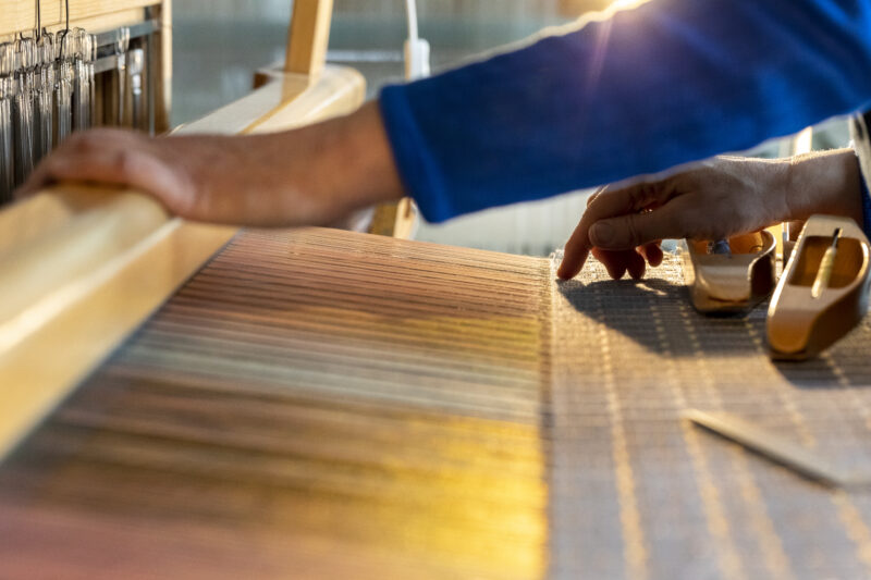 Hands working on a weaving loom, with colorful threads and wooden tools visible, illuminated by warm light.
