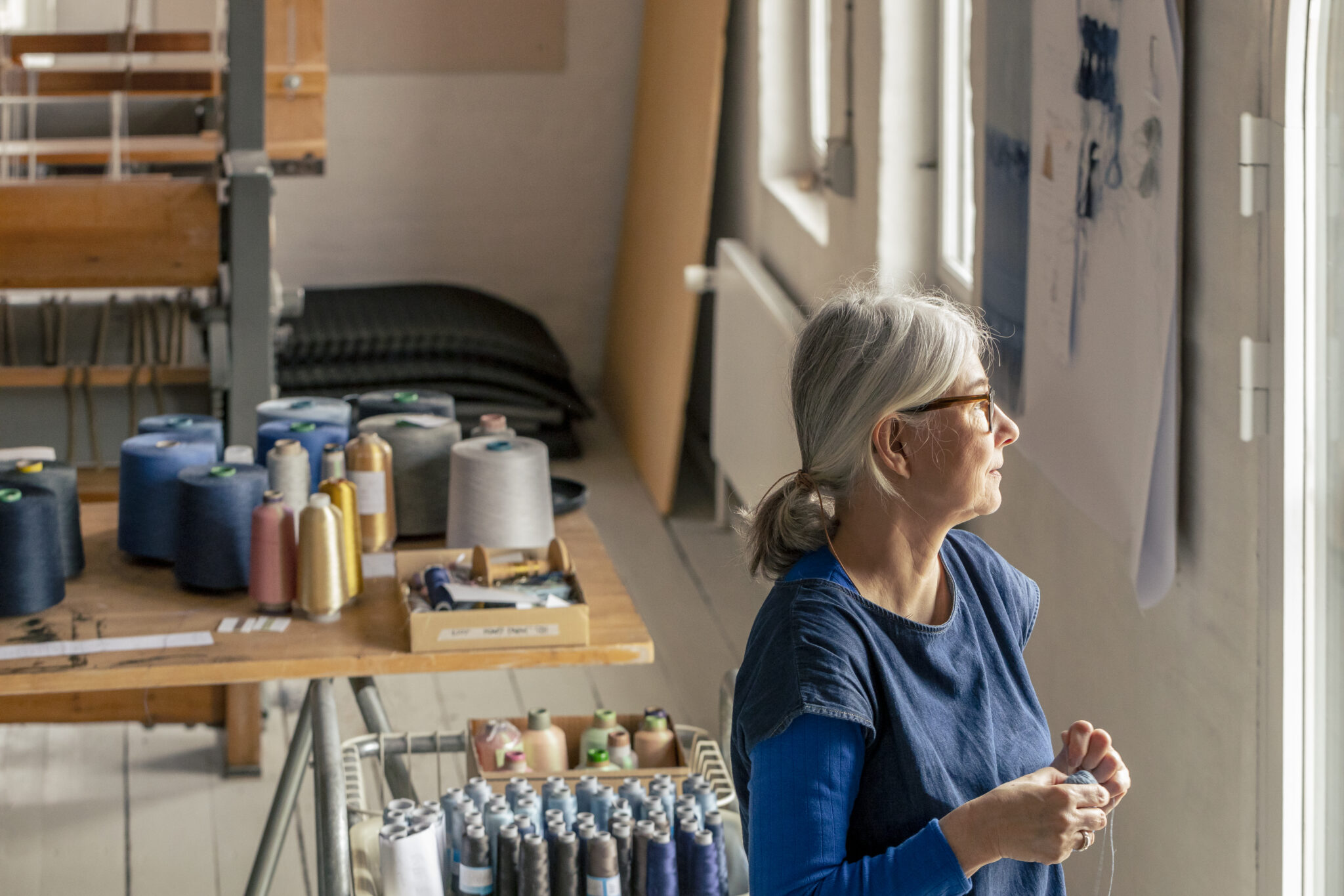 A woman with gray hair and glasses stands by a window in a textile workshop, surrounded by spools of thread.
