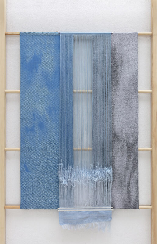 Textile artwork featuring three panels in varying shades of blue and gray, with vertical threads in the center.