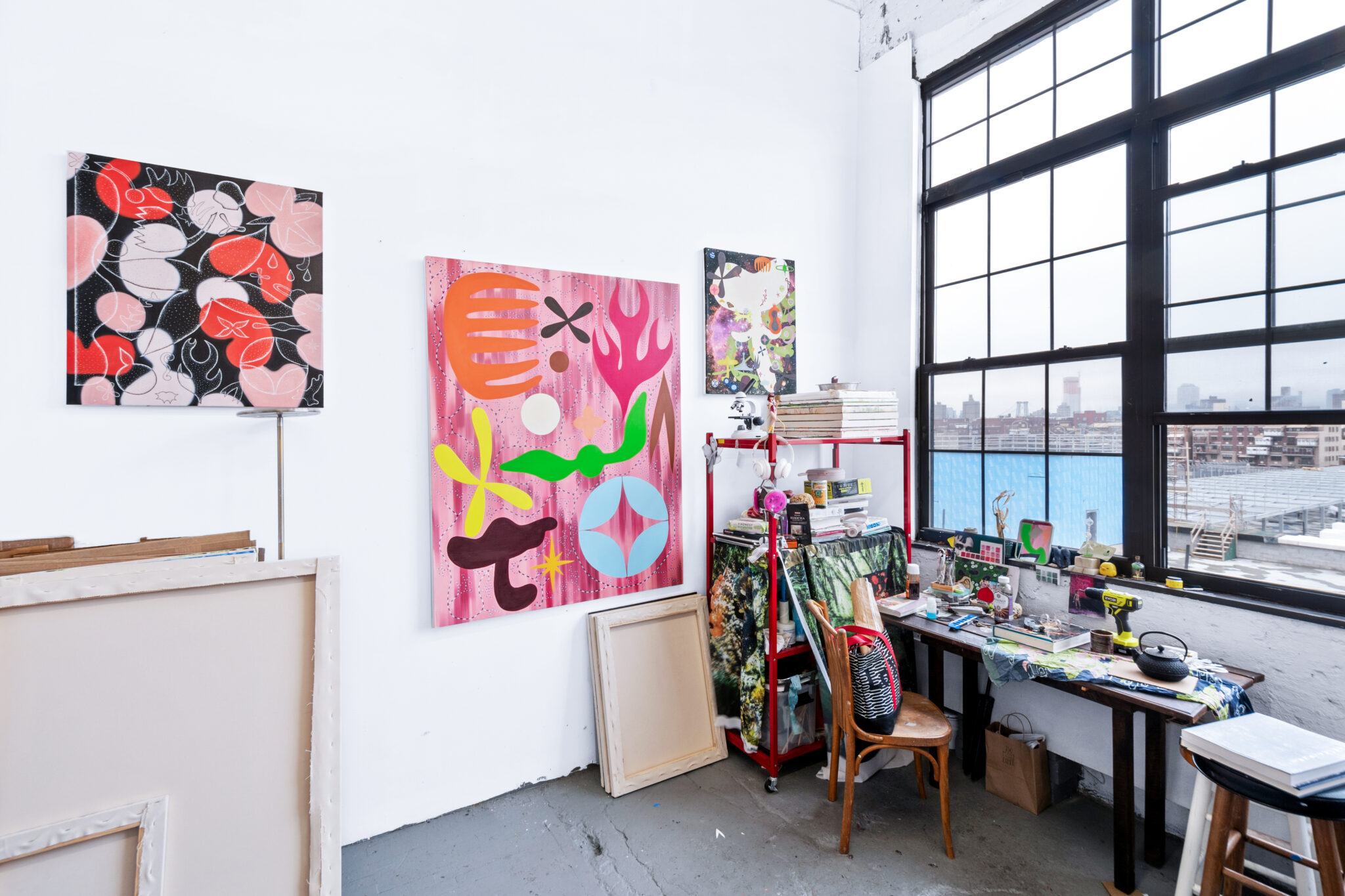 Colorful abstract artworks hang on a white wall in a bright studio, with a cluttered desk and large windows in the backgro...
