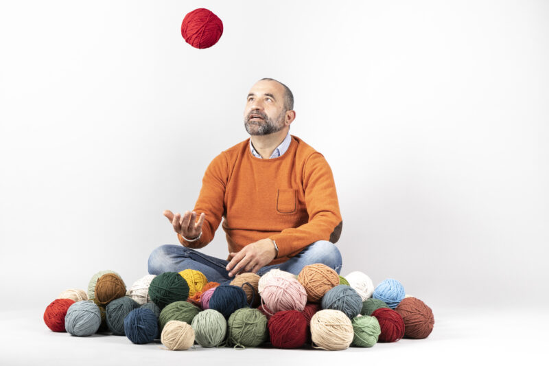 Man in an orange sweater juggling a red yarn ball, surrounded by various colored yarn balls on a white background.