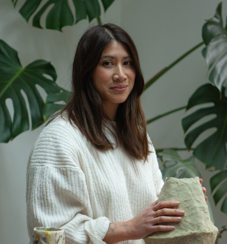 Woman in a white sweater poses with a clay vase among colorful pottery, surrounded by large green leaves.
