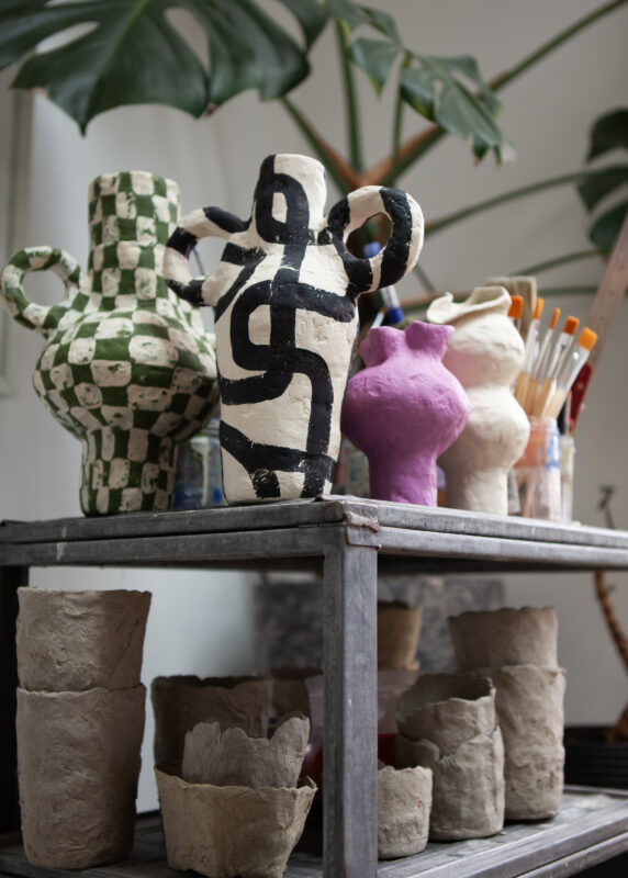 Colorful ceramic vases with various patterns on a metal shelf, surrounded by art supplies and plant leaves.