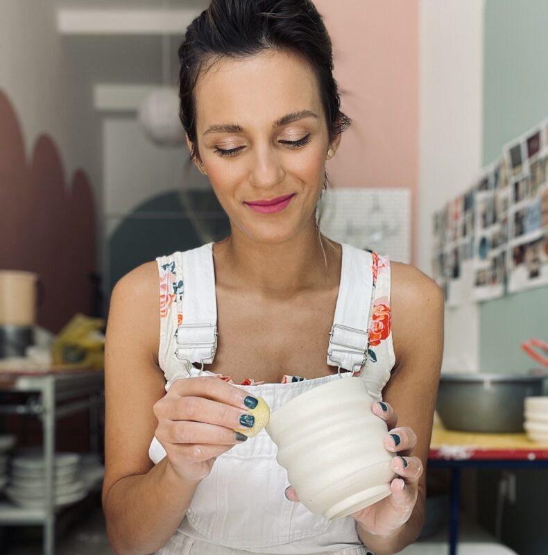 Woman in white overalls holding a ceramic bowl, smiling while crafting in a studio with pastel-colored walls.