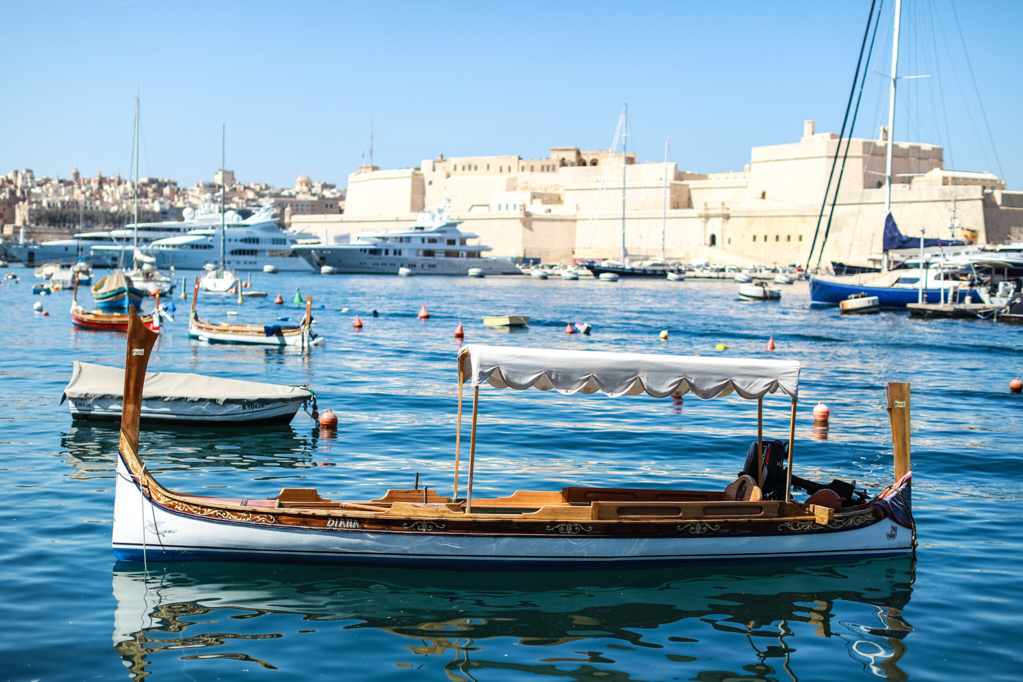A traditional luzzu boat floats in Grand Harbour, with historic buildings and other boats in the background.