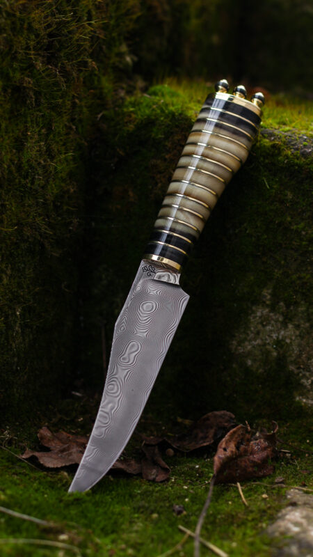 Damascus steel knife with a decorative handle, resting on moss and leaves against a stone background.