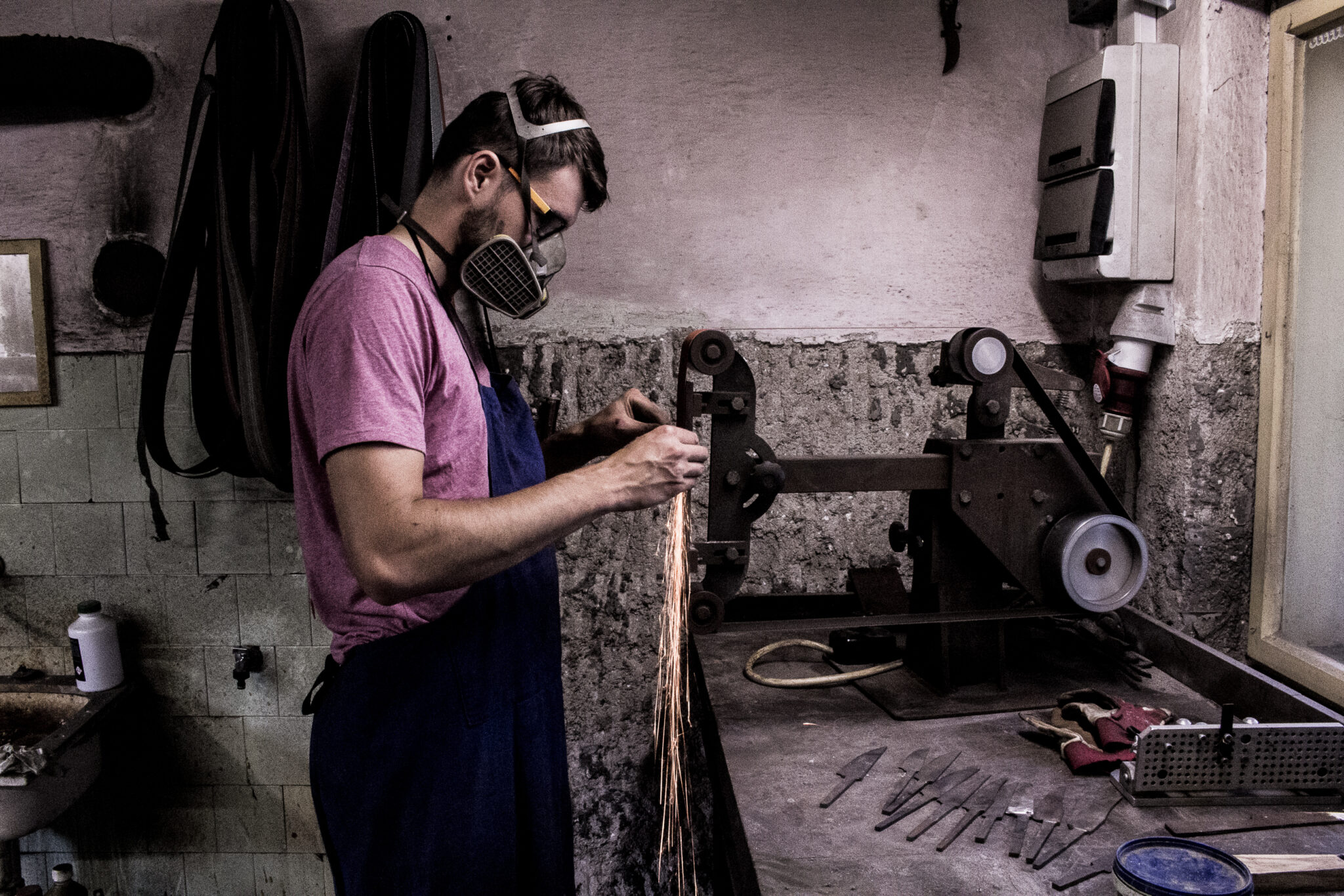 Man in a mask working on metal at a workshop, with tools and equipment visible on the table.