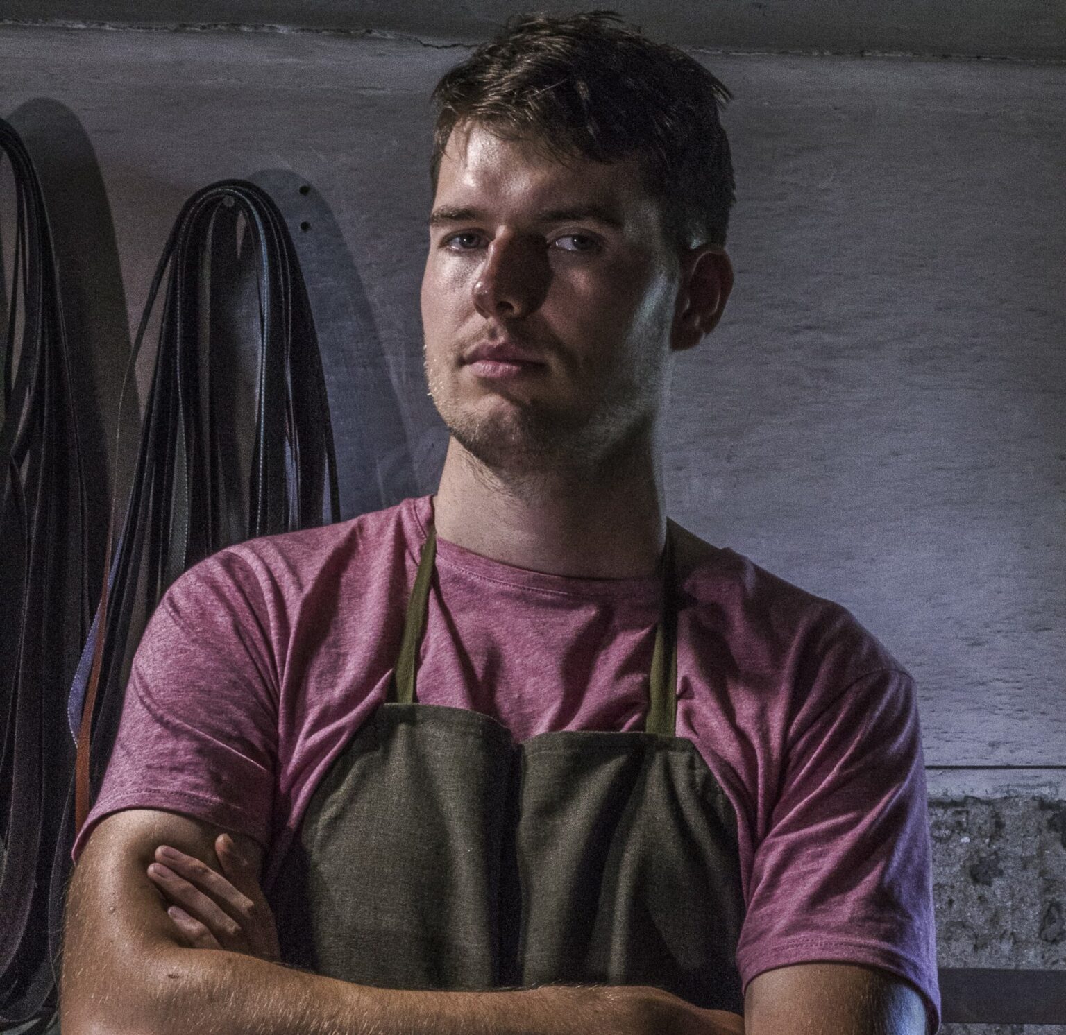 Young man with short hair, wearing a pink shirt and green apron, stands with arms crossed in a workshop setting.