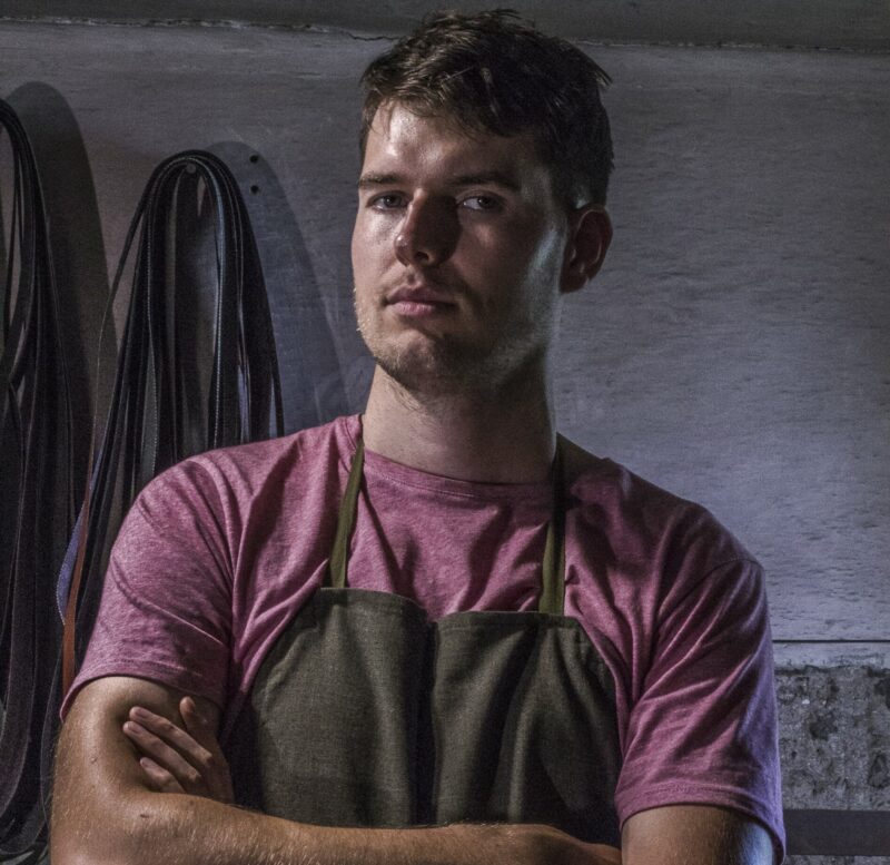 Young man with short hair, wearing a pink shirt and green apron, stands with arms crossed in a workshop setting.