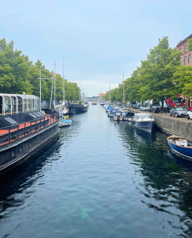 Calm waterway lined with boats and trees, leading to a distant harbor under a cloudy sky.