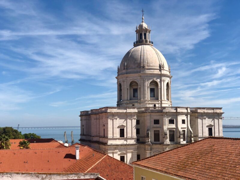 Lisbon skyline featuring the National Pantheon with red rooftops in the foreground and a blue sky.