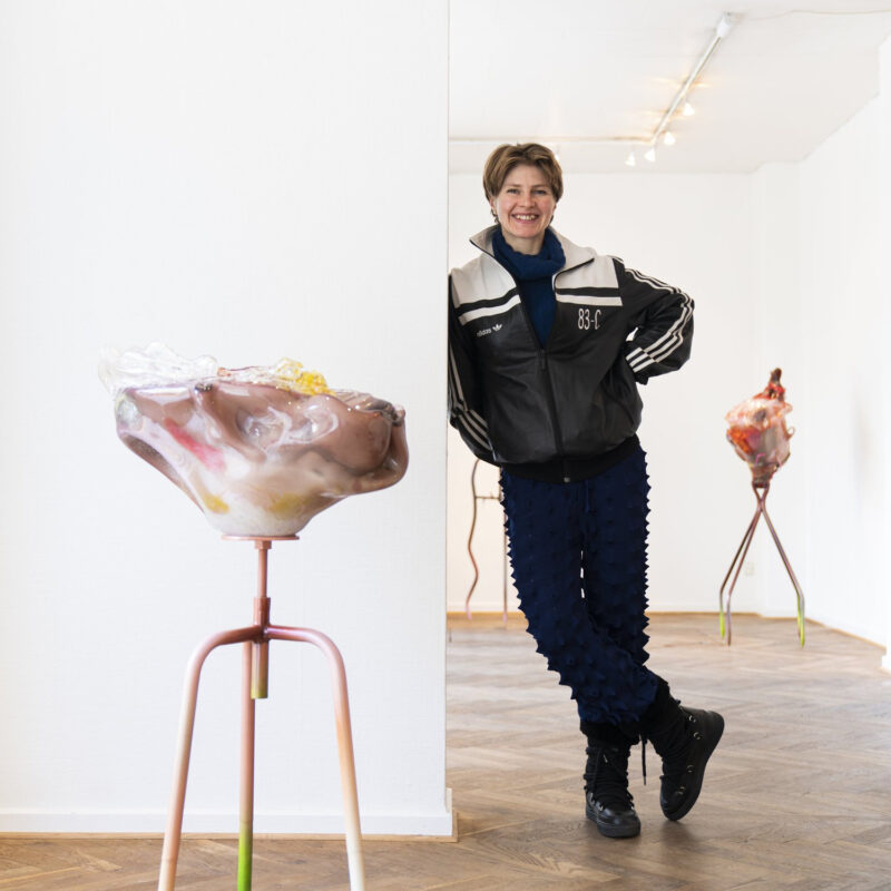 Woman in a black jacket and blue pants stands beside a wall, with abstract sculptures on stands in an art gallery.