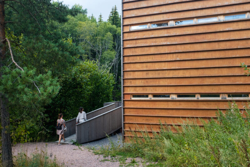 Two people walking on a wooden path towards a modern wooden building, surrounded by greenery at KWUM Museum, Fiskars.