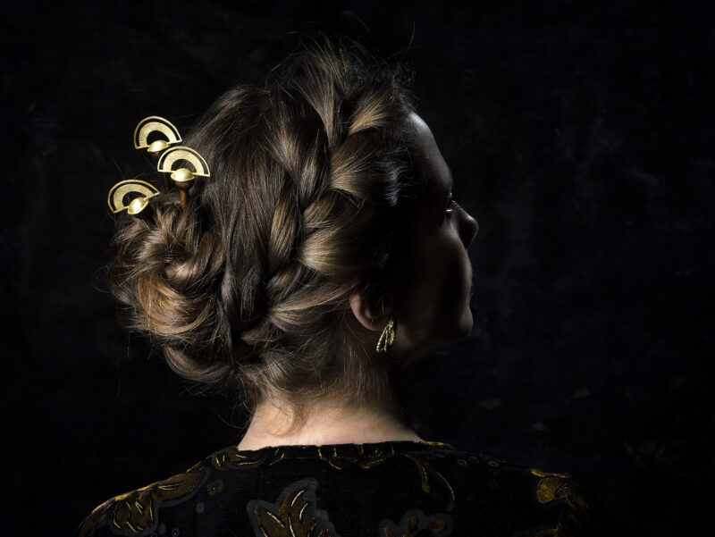 Woman with braided hair adorned with gold hairpins, viewed from the side against a dark background.
