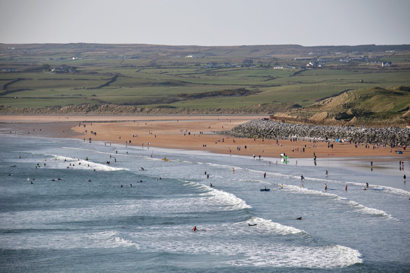 Lahinch beach in Co Clare, featuring surfers in the waves and a sandy shoreline, surrounded by green hills.