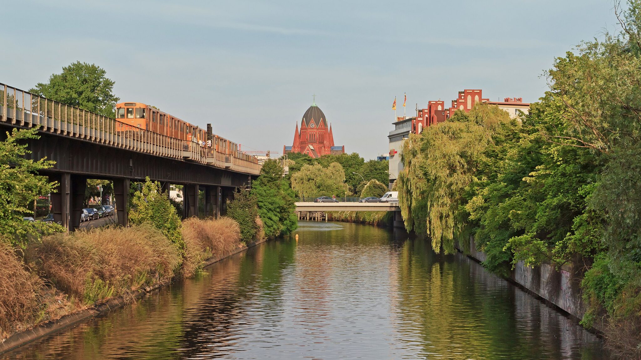View of Landwehrkanal in B-Kreuzberg, Berlin, with a train on an elevated track and greenery along the waterway.
