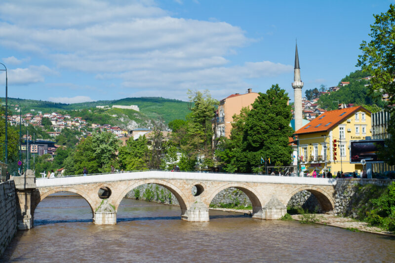 Latin Bridge in Sarajevo spans a river, surrounded by greenery and buildings, with a minaret visible in the background.