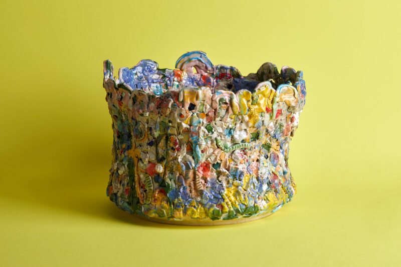 Colorful ceramic vessel resembling a human body, textured and abstract, set against a bright yellow background.