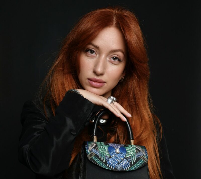 Liana Tashchyan poses with a small black handbag featuring a colorful floral design against a dark background.