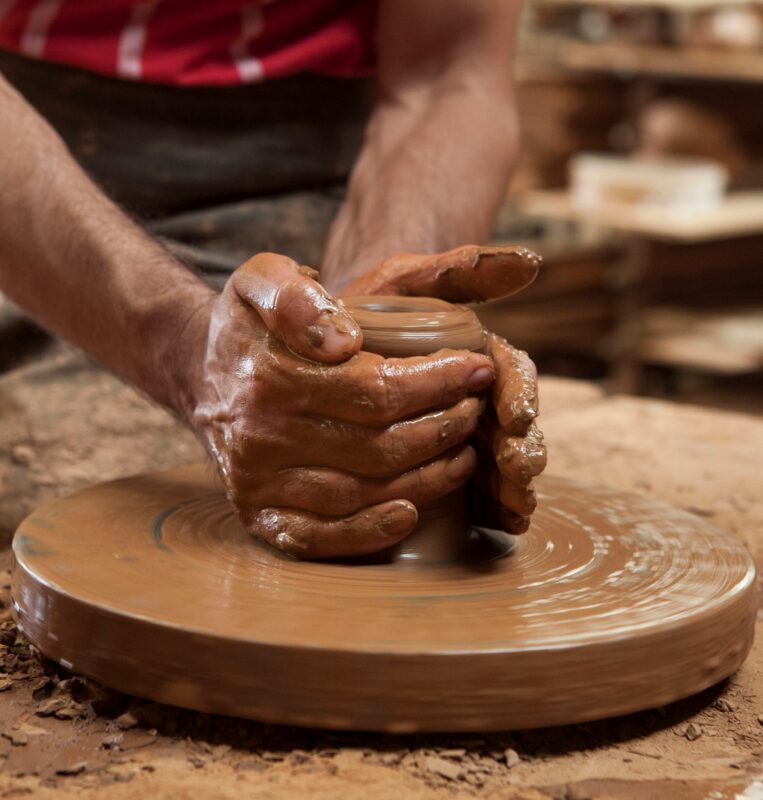 Hands shaping a clay pot on a pottery wheel in a workshop setting, with clay and tools visible around.
