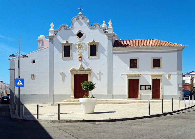 White building with ornate facade and red doors, surrounded by a circular plaza in Loule, Portugal.