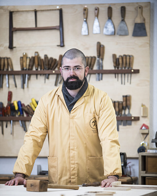 Man in a yellow work jacket stands at a wooden table in a workshop, tools hanging on the wall behind him.