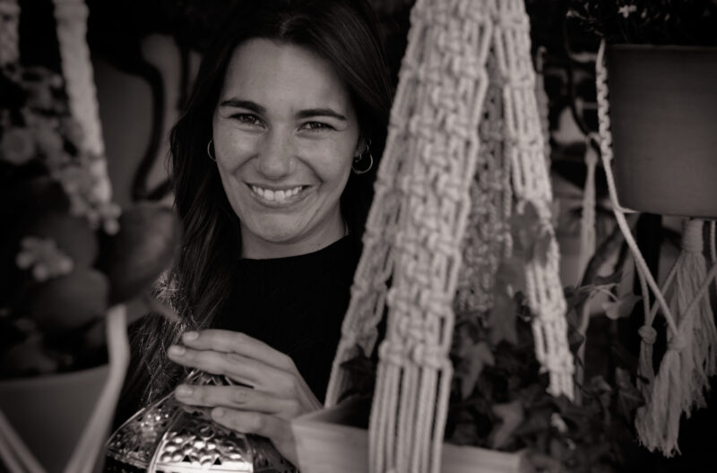 Smiling woman holding a decorative lantern, surrounded by hanging plants and macramé decor.