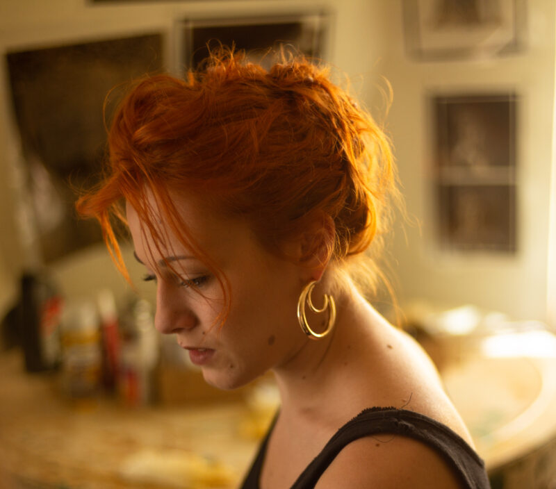 Portrait of a woman with orange hair, wearing large hoop earrings, gazing down thoughtfully in a softly lit room.