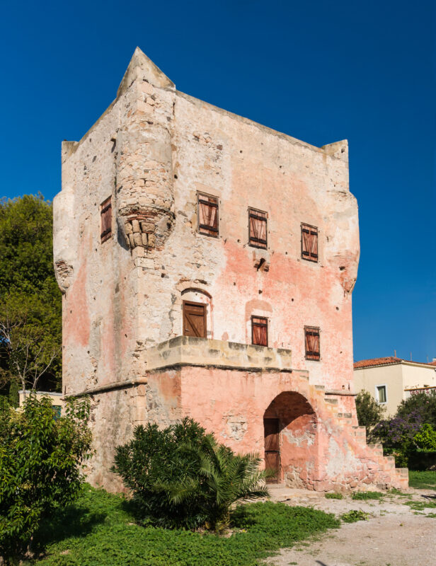 Markellos Tower on Aegina Island, featuring a weathered pink facade and wooden windows against a clear blue sky.