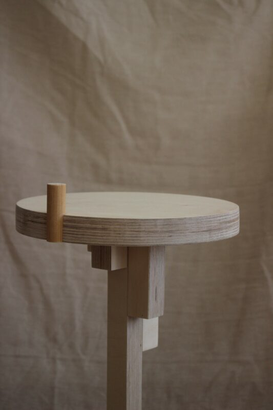 Minimalist round table with a light wood finish and a cylindrical leg, set against a neutral fabric background.