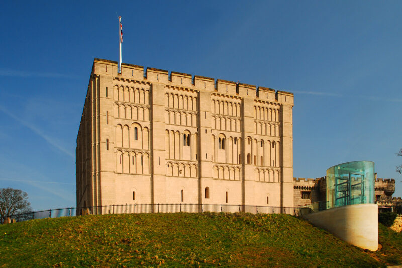 Norwich Castle keep, a large stone structure with a flag, set on a grassy hill under a clear blue sky.