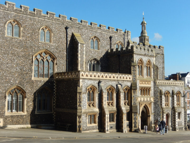 Norwich Guildhall's flint and stone façade, featuring ornate windows and a tower, under a clear blue sky.
