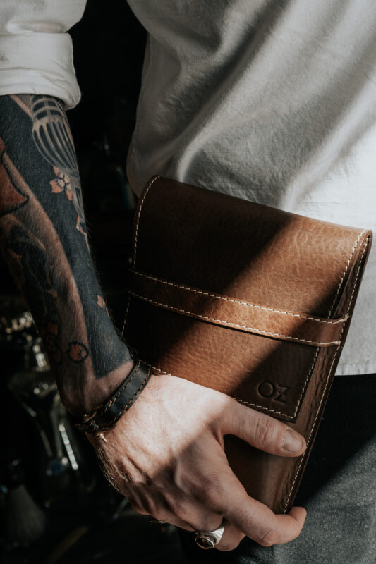Brown leather bag held by a tattooed arm, with sunlight casting shadows, titled "OLHA RUCCYA 1" by Olha Ruccya.