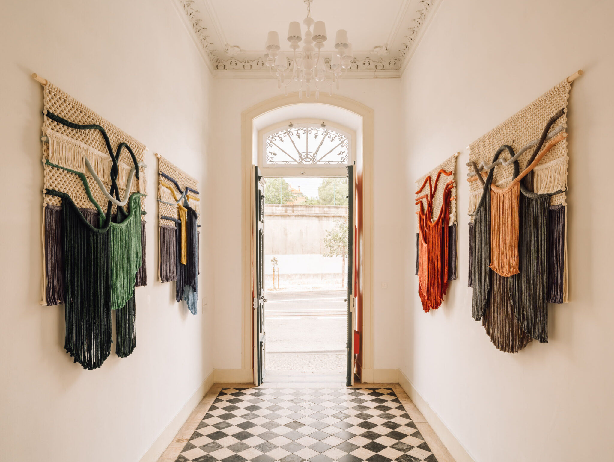 Colorful wall hangings by Vasco Águas displayed in a bright hallway with a checkered floor and open doors.