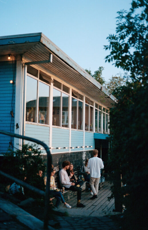 People enjoying food outside a blue building with large windows at sunset, surrounded by greenery.