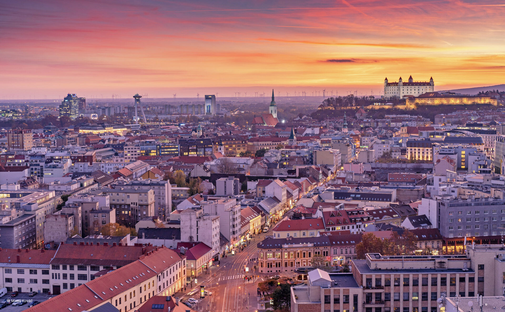 Sunset over Bratislava, showcasing the cityscape with buildings, streets, and the castle in the distance.