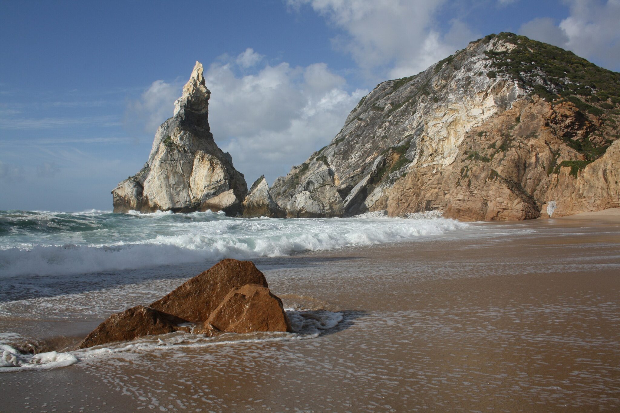 Rocky cliffs and a distinctive spire rise above Praia da Ursa beach, with waves lapping at the sandy shore.