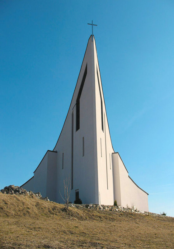Modern church with a tall, pointed roof against a clear blue sky, surrounded by grassy terrain.