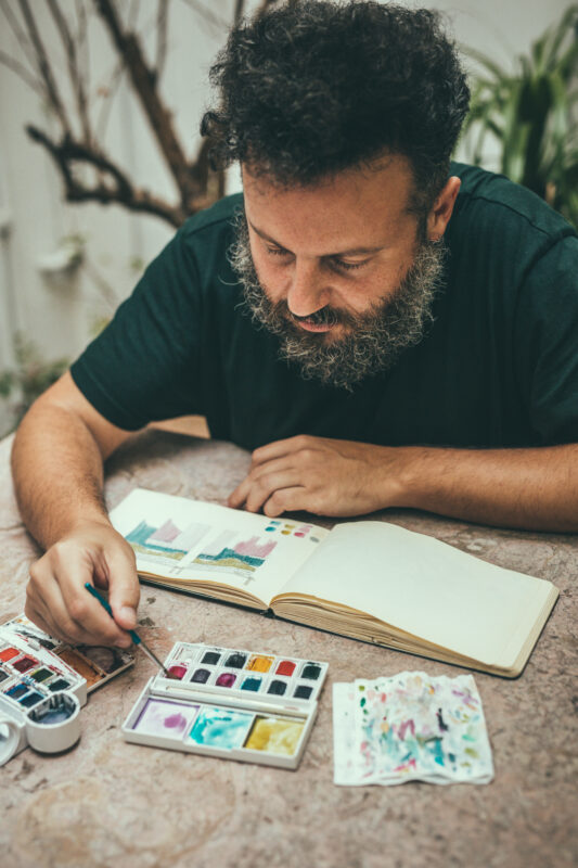 Artist Arlindo Camacho sketching in a notebook with watercolor paints on a table, surrounded by plants.