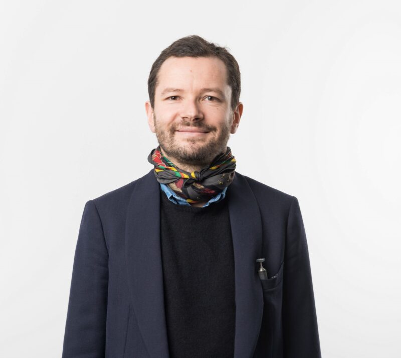 Man in a dark blazer and black sweater, wearing a colorful scarf, smiling against a white background.