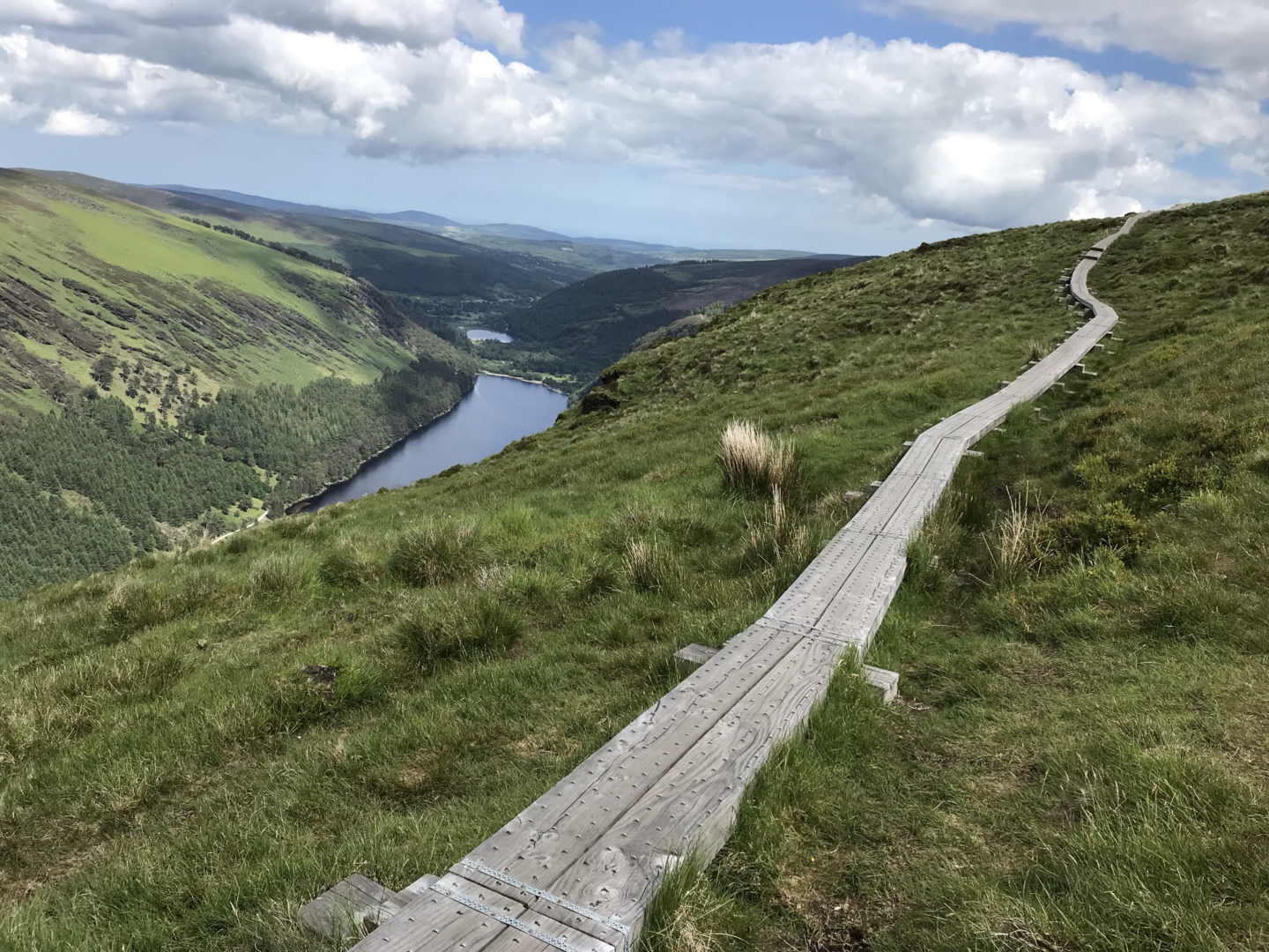 Lush green landscape with a wooden path leading to a lake in the Wicklow Mountains, under a partly cloudy sky.