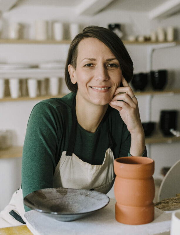 Tatjana Kovačević-Vidović at TAKO pottery studio, smiling with pottery pieces and tools on a worktable.