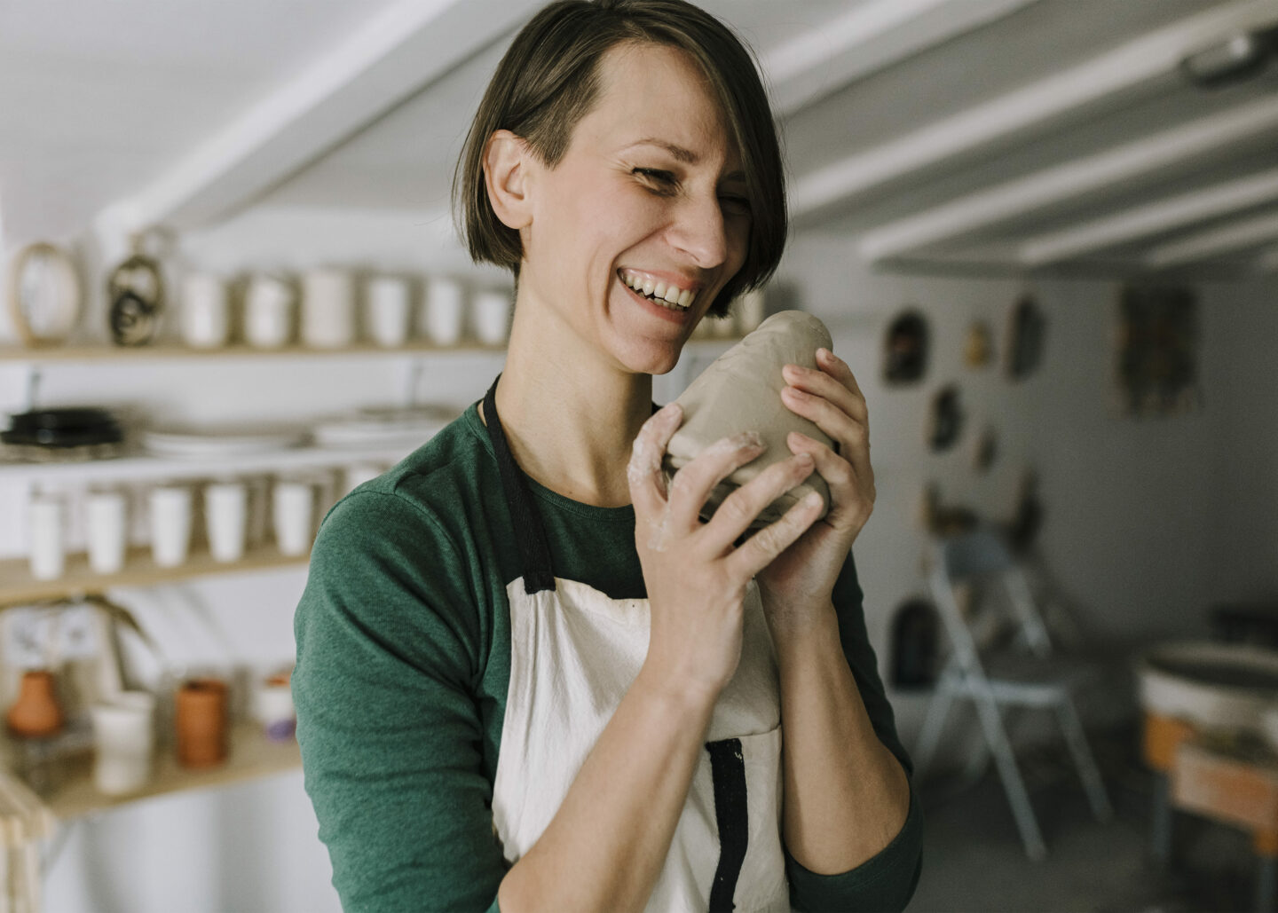 Woman in a pottery studio joyfully holds a lump of clay, surrounded by pottery on shelves.
