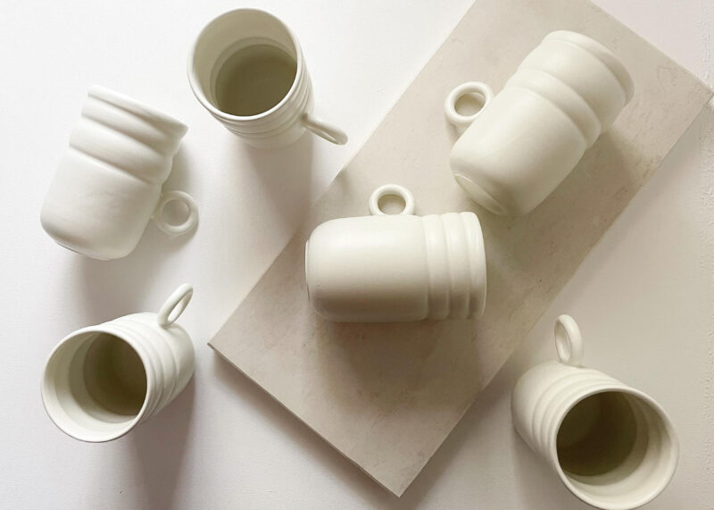 White ceramic mugs in various positions on a light surface, showcasing their tall, cylindrical shapes and handles.