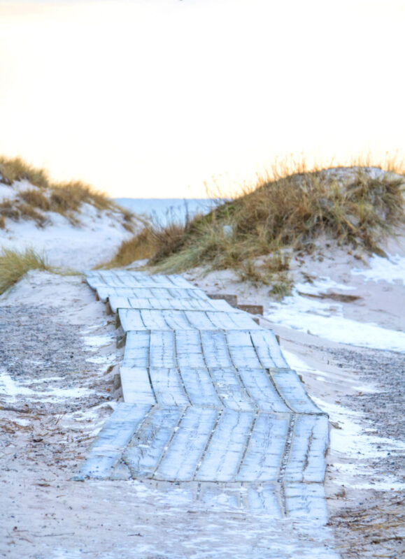 Wooden walkway leading through sandy dunes at Sandhammaren beach, with grass and soft light in the background.