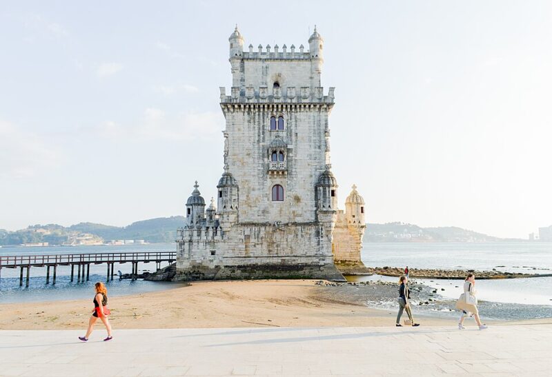 Torre de Belém, a historic tower in Lisbon, with people walking along the sandy shore and a pier in the background.